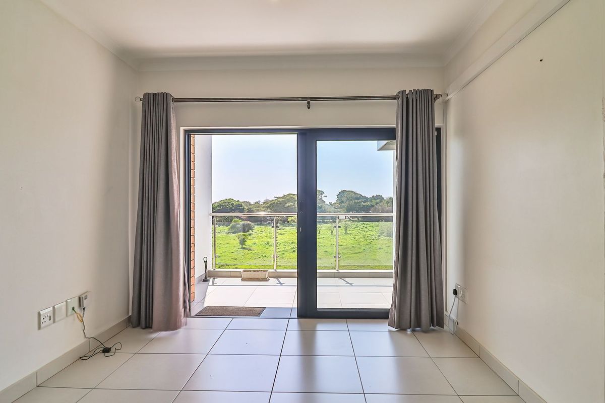 A bright, empty living area featuring large sliding glass doors that open onto a balcony with scenic green views and a modern glass balustrade.
