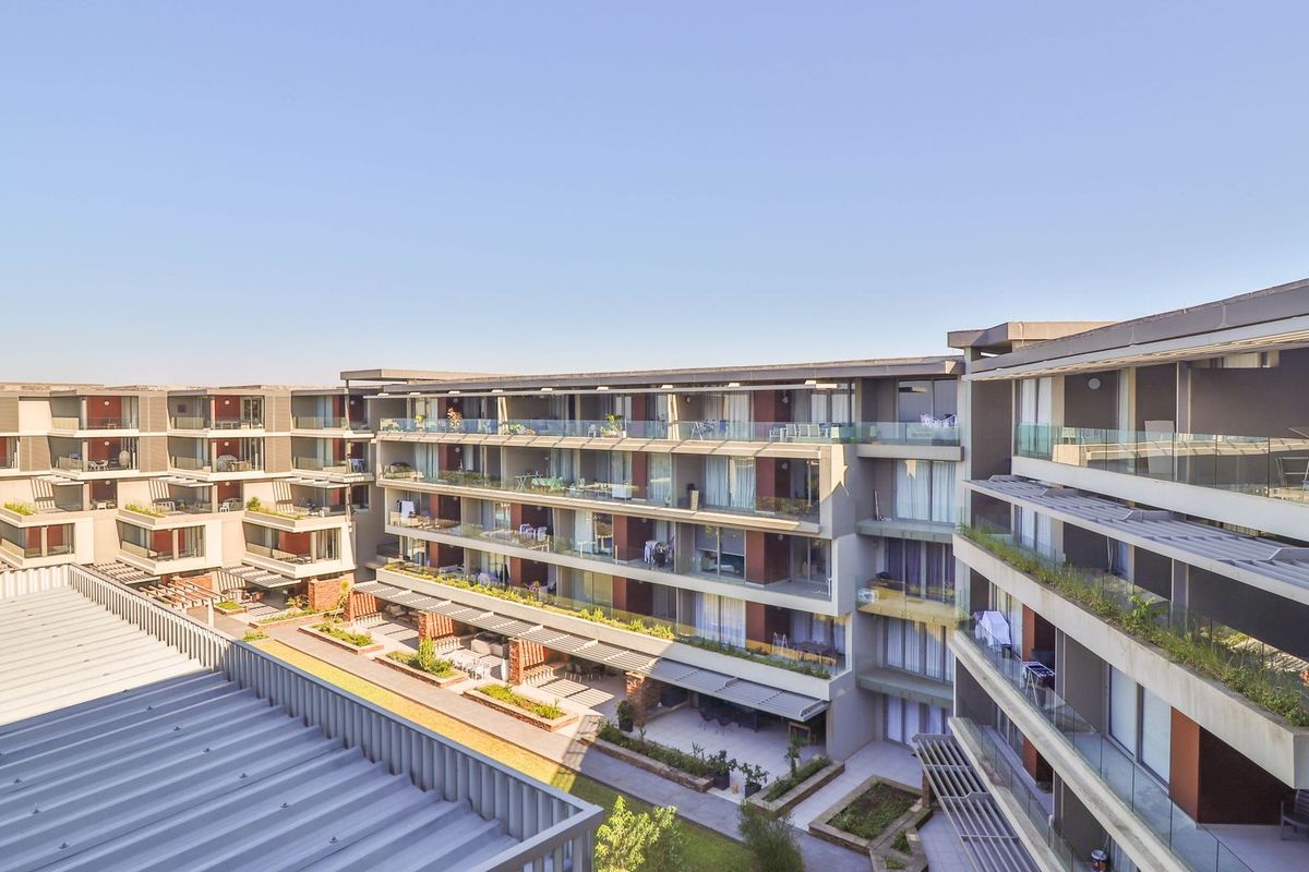 An elevated aerial view of a modern apartment complex featuring multi-level balconies, glass railings, and landscaped ground-level patio areas.