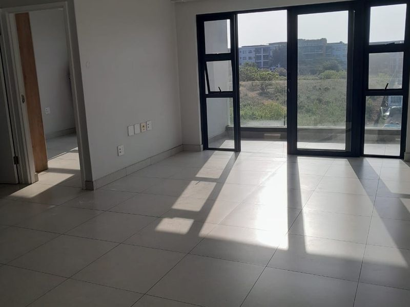 A bright, open-plan living area featuring large black-framed glass sliding doors that open onto a balcony, with neutral tiled flooring throughout.