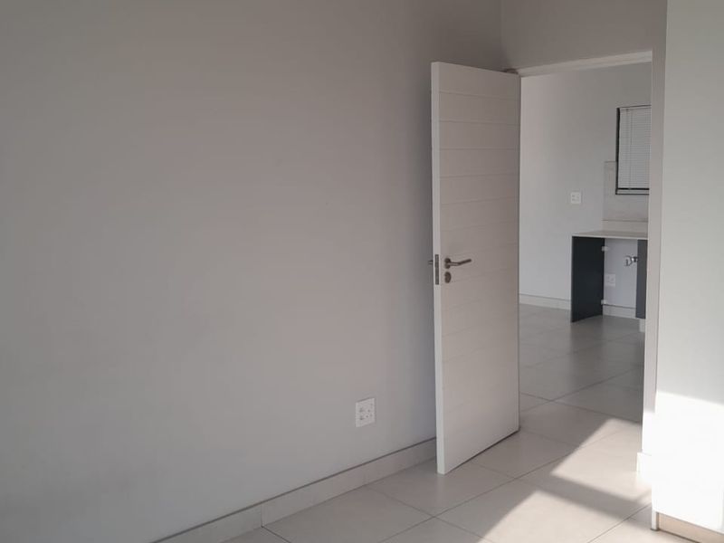 A bright, empty bedroom featuring neutral tiled flooring and a doorway leading to an adjacent kitchen area.