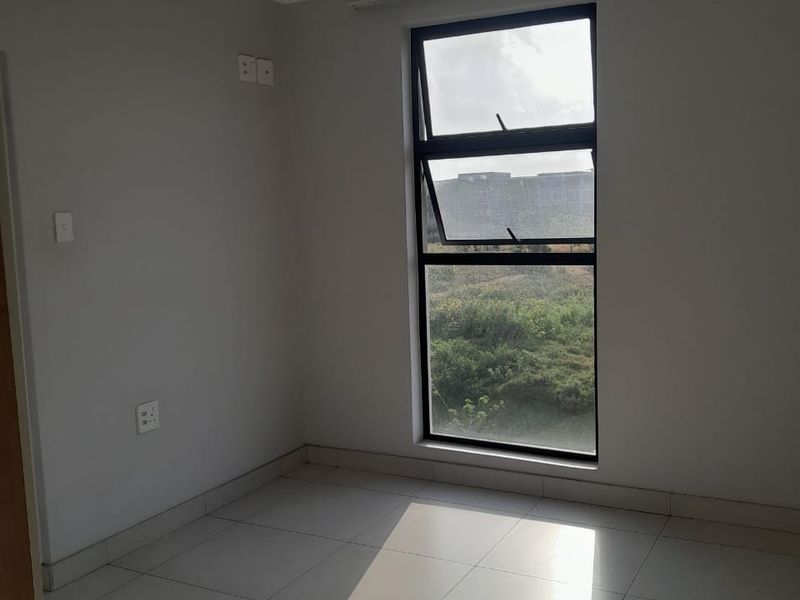 A bright, empty bedroom featuring neutral tiled flooring, a large vertical window with natural light, and a simple ceiling-mounted light fixture.