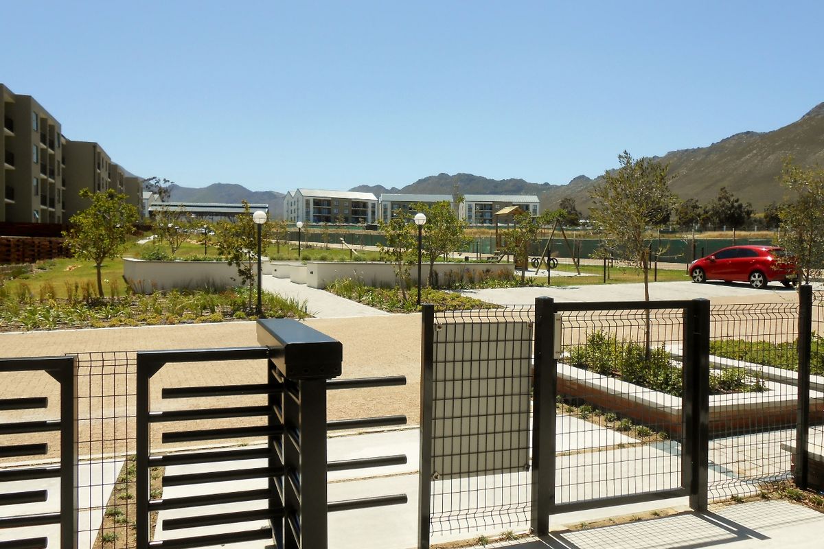 A view of the communal landscaped garden area and paved pathways within the apartment complex, featuring perimeter fencing and mountain views.