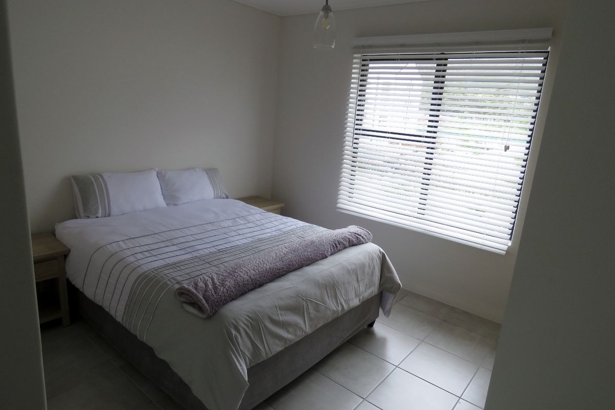 A well-lit bedroom featuring neutral tiled flooring, a large window with venetian blinds, and a modern pendant light fixture.