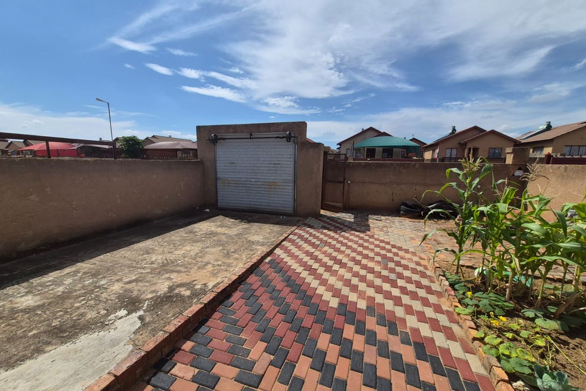 A rear exterior view featuring a paved walkway, a concrete area, and a secure boundary wall with a roller garage door.