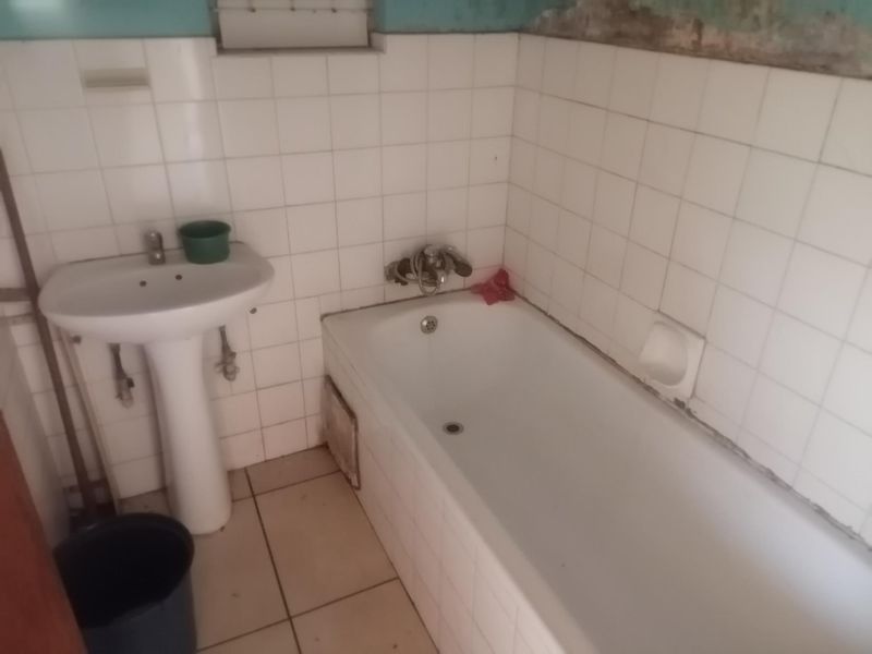 A dated bathroom featuring a pedestal basin and bathtub with white wall tiling, requiring renovation to address damaged wall finishes and general wear.