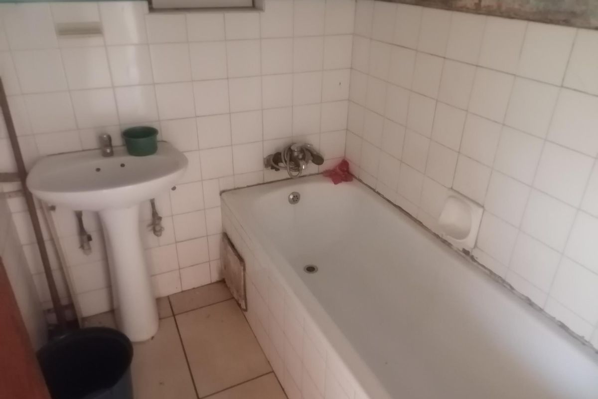 A dated bathroom featuring a pedestal basin and bathtub with white wall tiling, requiring renovation to address damaged wall finishes and general wear.