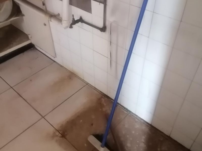 A kitchen area featuring tiled walls and flooring with exposed plumbing and wall-mounted tap, requiring maintenance and cosmetic updates.