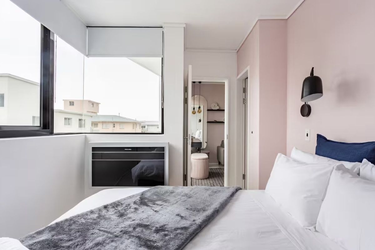 A bright bedroom featuring large corner windows, built-in air conditioning unit, and direct access to an adjoining room.