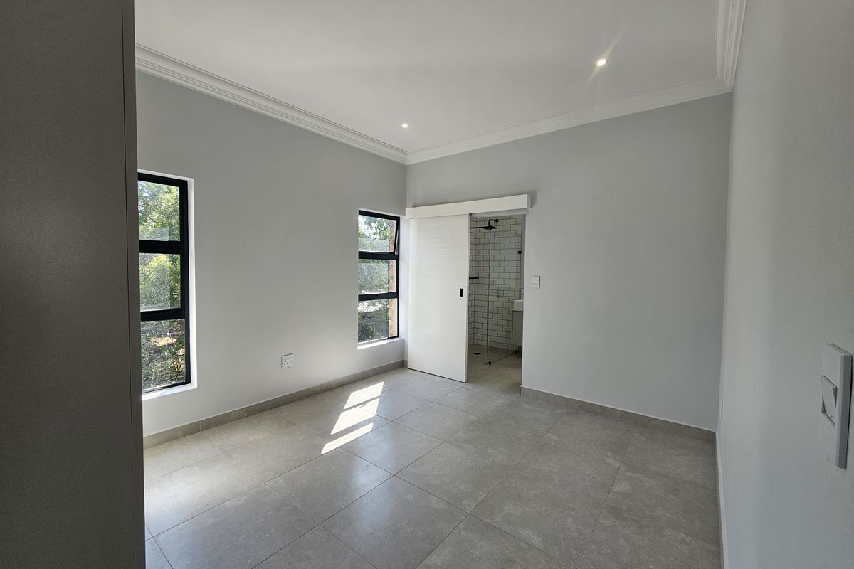 A bright, modern bedroom featuring neutral tiled flooring, recessed ceiling lighting, and a sliding door leading to an en-suite bathroom.