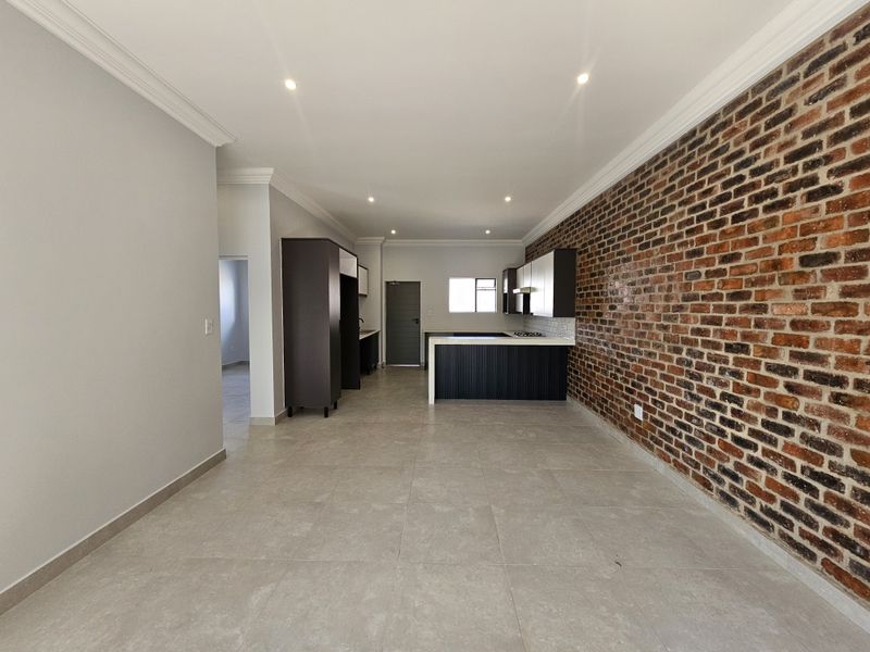 Open plan kitchen and living area featuring a striking exposed brick feature wall, modern cabinetry, and neutral floor tiling throughout.