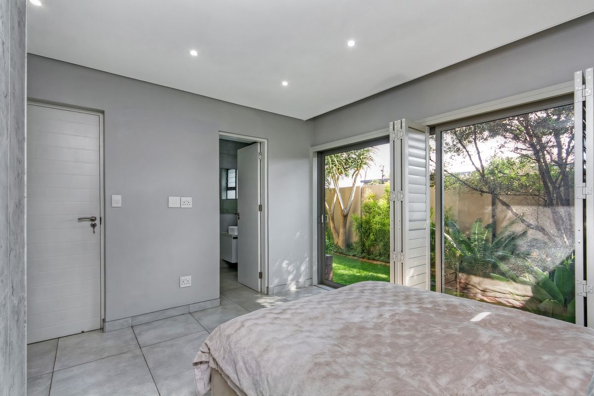 Modern bedroom featuring tiled floors, recessed lighting, and large folding shutters opening to a private garden view, with direct access to an en-suite bathroom.