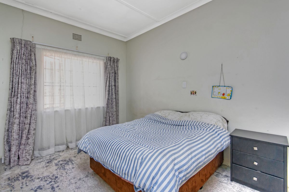 A bedroom featuring a window with curtains, ceiling cornice, and ventilation grille. The room shows signs of wear including wall cracks and scuffed paint.