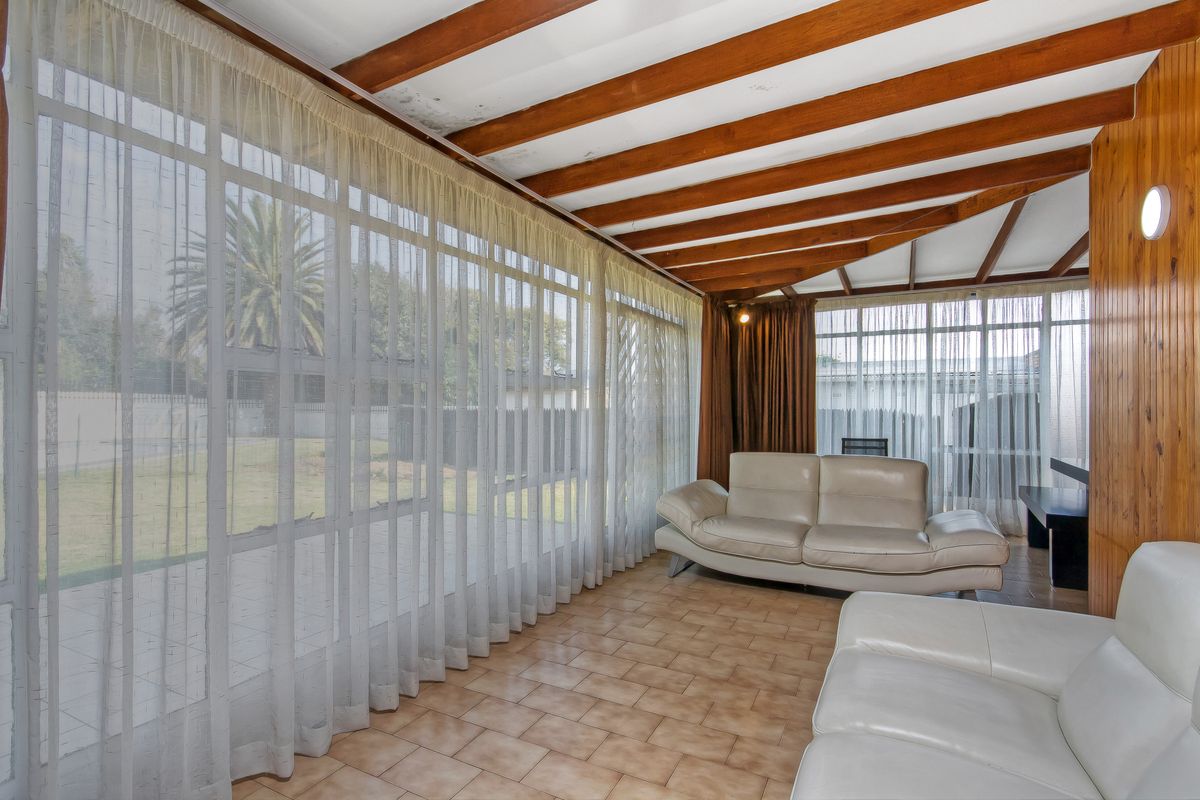 Sun-filled living room featuring exposed timber ceiling beams, wood wall paneling, and large windows overlooking the garden.