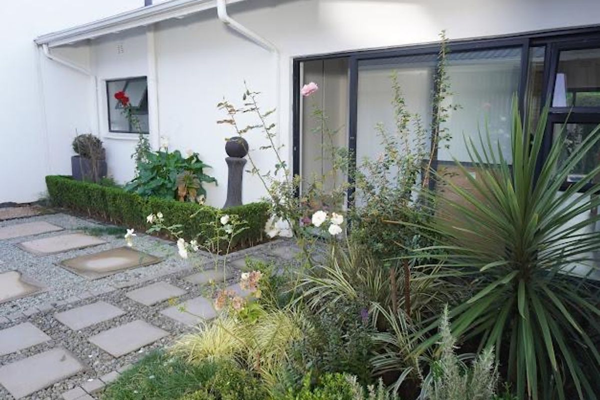 A well-maintained rear garden area featuring paved stepping stones, gravel landscaping, and established garden beds adjacent to the building's sliding glass doors.