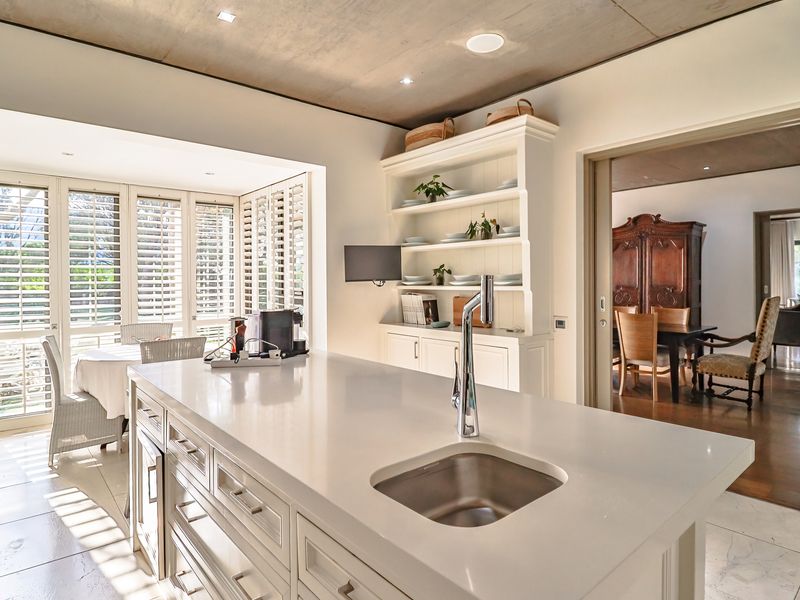 Modern kitchen featuring a central island with a stone countertop, integrated sink, and custom cabinetry, opening into a bright dining area with plantation shutters.
