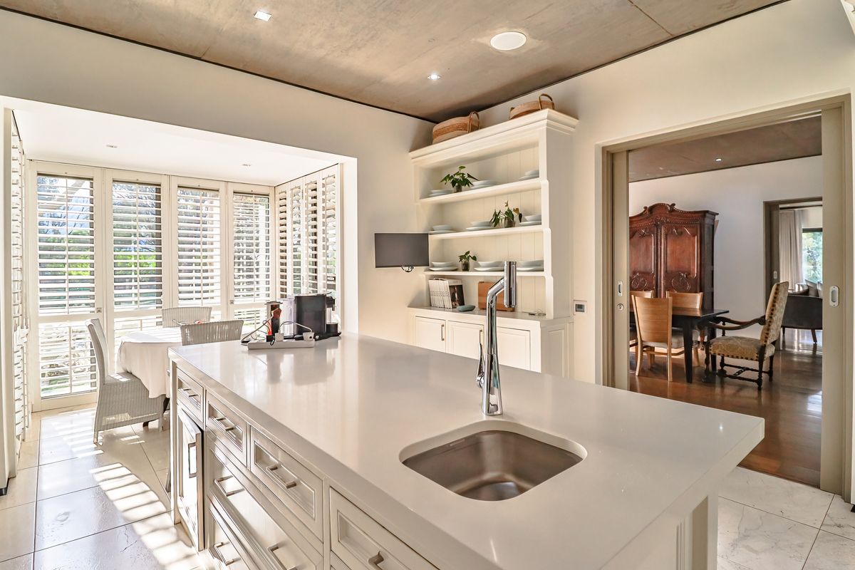 Modern kitchen featuring a central island with a stone countertop, integrated sink, and custom cabinetry, opening into a bright dining area with plantation shutters.