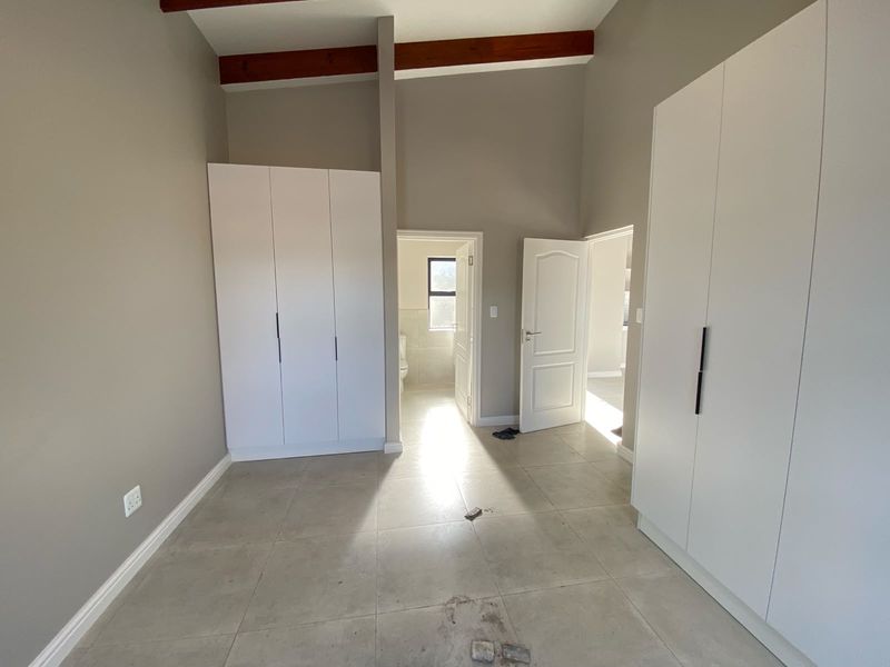 A spacious, empty bedroom featuring modern built-in cupboards, neutral tiled flooring, and exposed timber ceiling beams, with direct access to an en-suite bathroom.