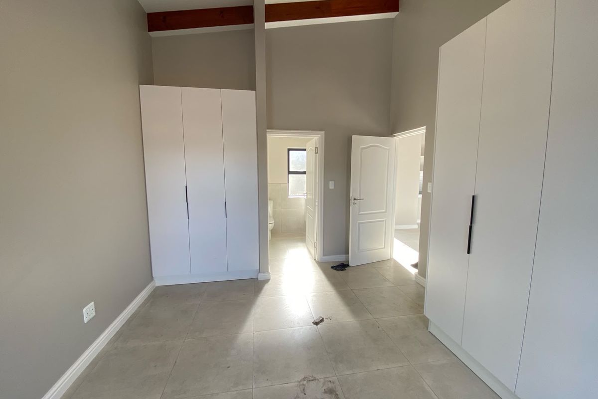 A spacious, empty bedroom featuring modern built-in cupboards, neutral tiled flooring, and exposed timber ceiling beams, with direct access to an en-suite bathroom.