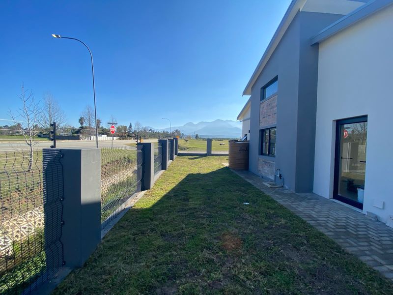 Side view of the property featuring a paved walkway, mesh perimeter fencing, water tank, and mountain views in the background.