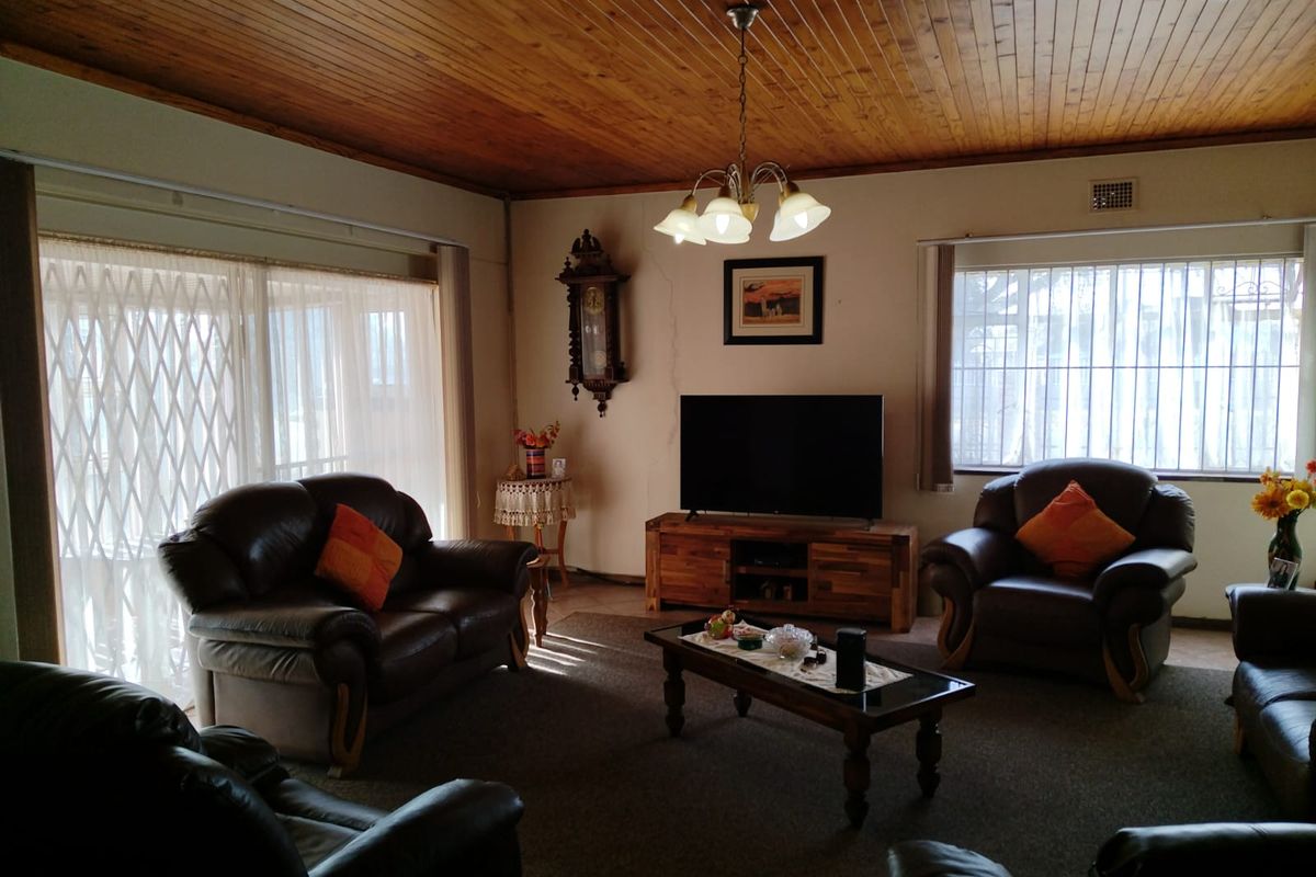 A spacious living room featuring a traditional timber tongue and groove ceiling, large windows with security bars, and a sliding door with a security gate.