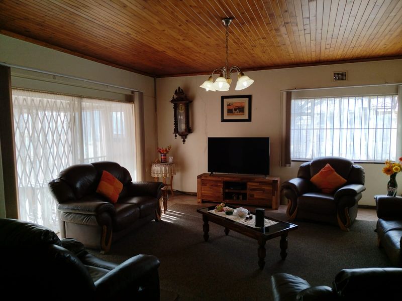 A spacious living room featuring a traditional timber tongue and groove ceiling, large windows with security bars, and a sliding door with a security gate.