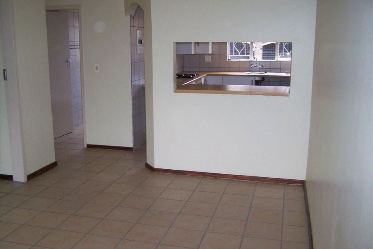 An empty living area featuring tiled flooring and a serving hatch opening into the kitchen, with an adjacent hallway leading to other rooms.