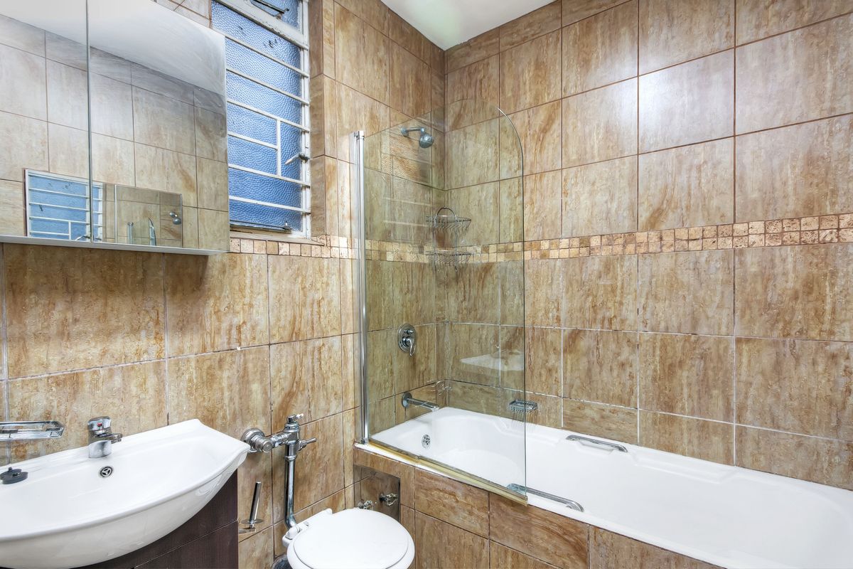 A compact bathroom featuring a bathtub with a glass shower screen, wall-mounted basin, and full-height brown ceramic wall tiles.