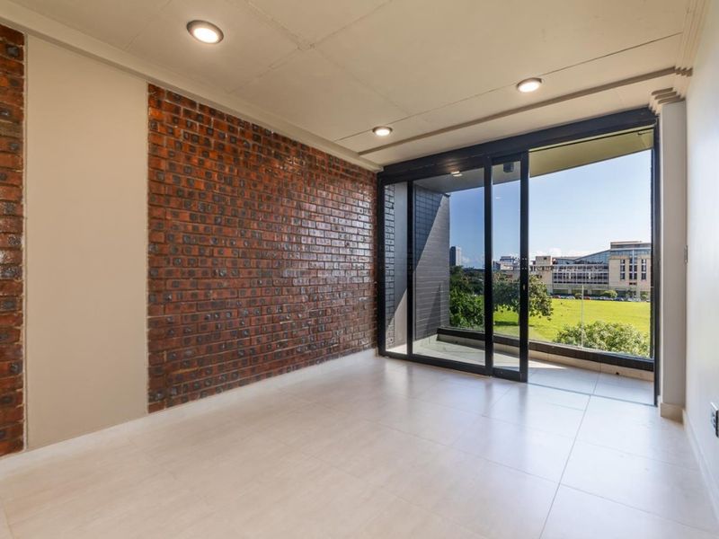 A bright, empty living area featuring a striking exposed brick feature wall, tiled flooring, recessed lighting, and large sliding glass doors opening to a balcony.