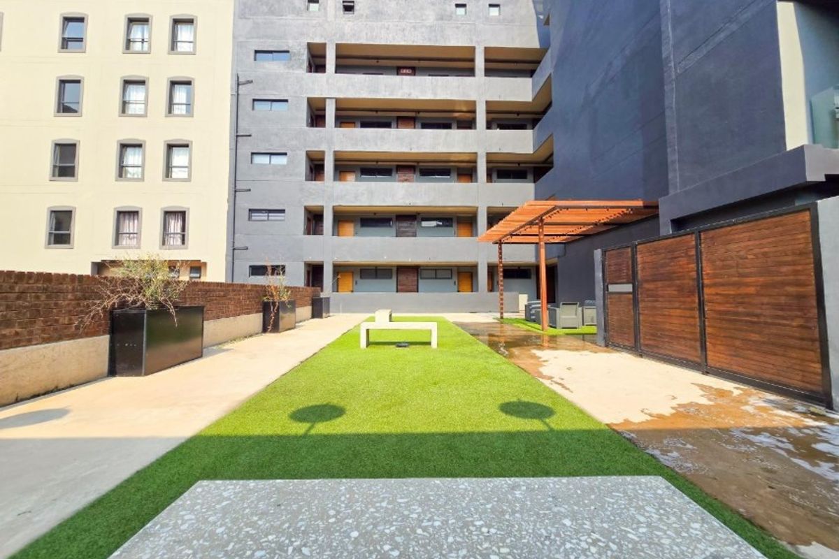 A communal outdoor courtyard area featuring artificial lawn, a wooden pergola, and brick boundary walls within an apartment complex.