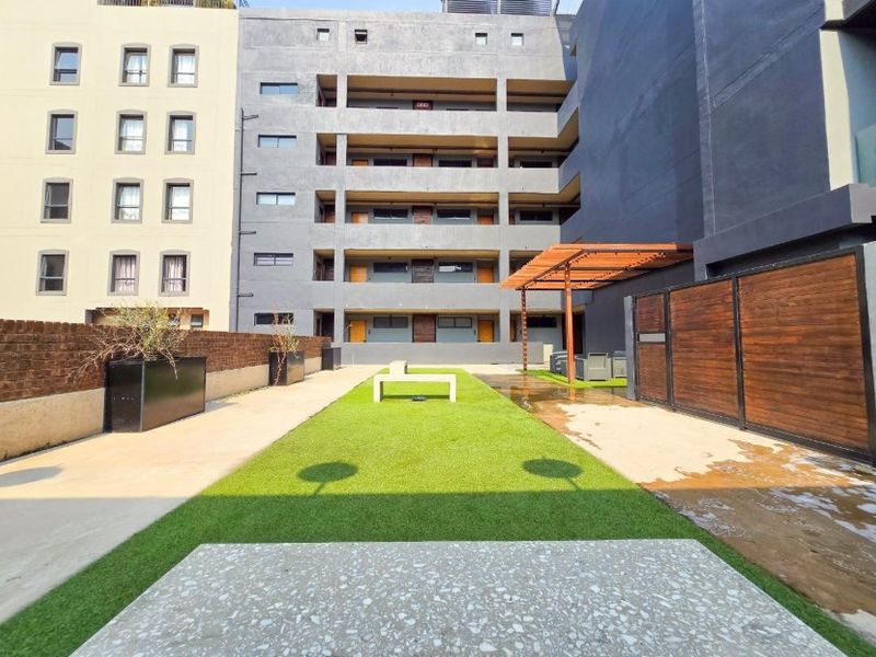A communal outdoor courtyard area featuring artificial lawn, a wooden pergola, and brick boundary walls within an apartment complex.