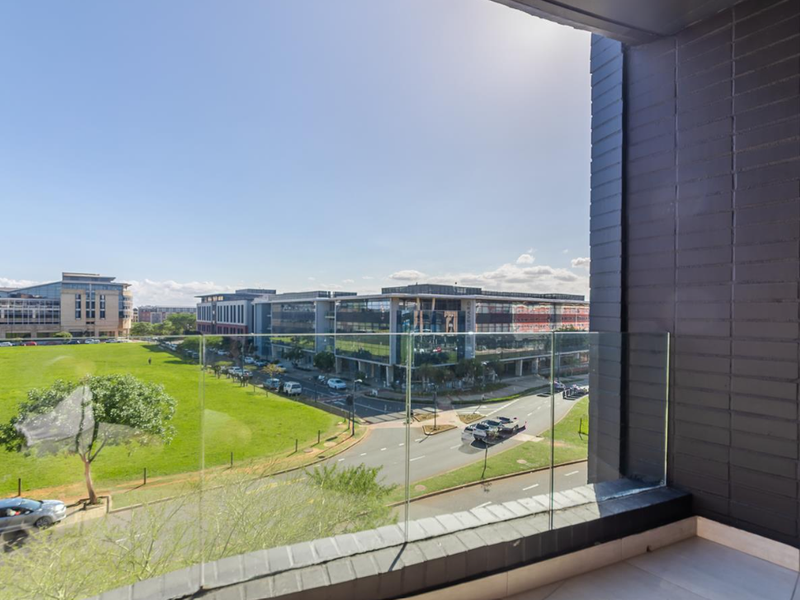 A modern balcony featuring a glass balustrade and tiled flooring, offering an elevated view of the surrounding urban landscape and green space.
