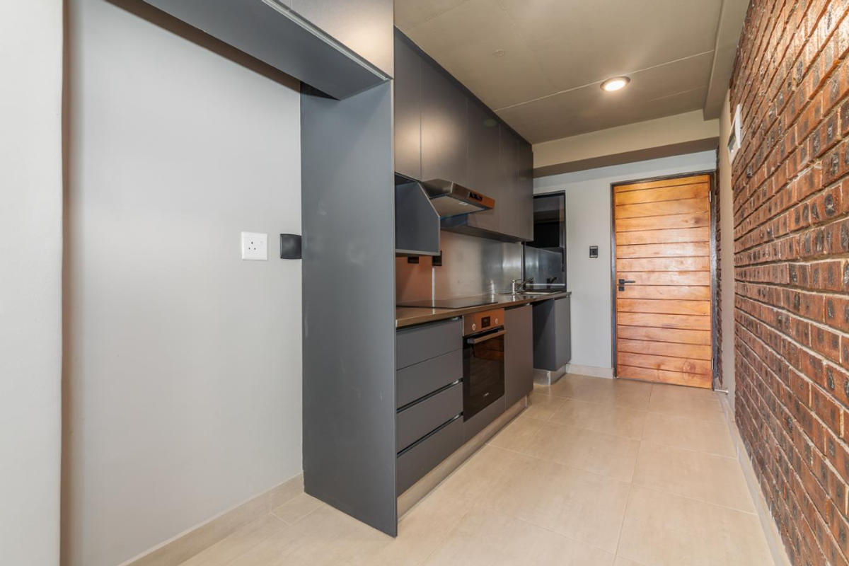 Modern galley-style kitchen featuring dark cabinetry, integrated oven and hob, stainless steel backsplash, and a striking exposed brick feature wall.