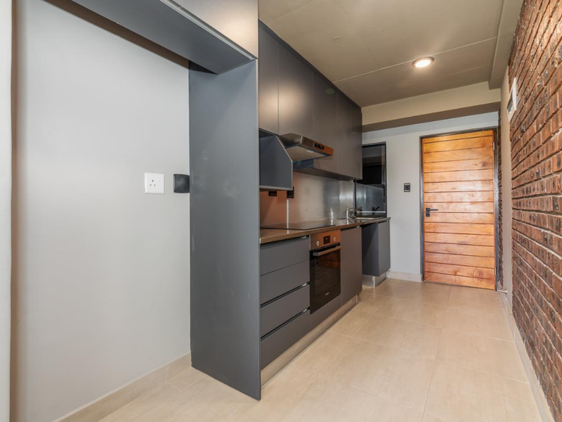 Modern galley-style kitchen featuring dark cabinetry, integrated oven and hob, stainless steel backsplash, and a striking exposed brick feature wall.