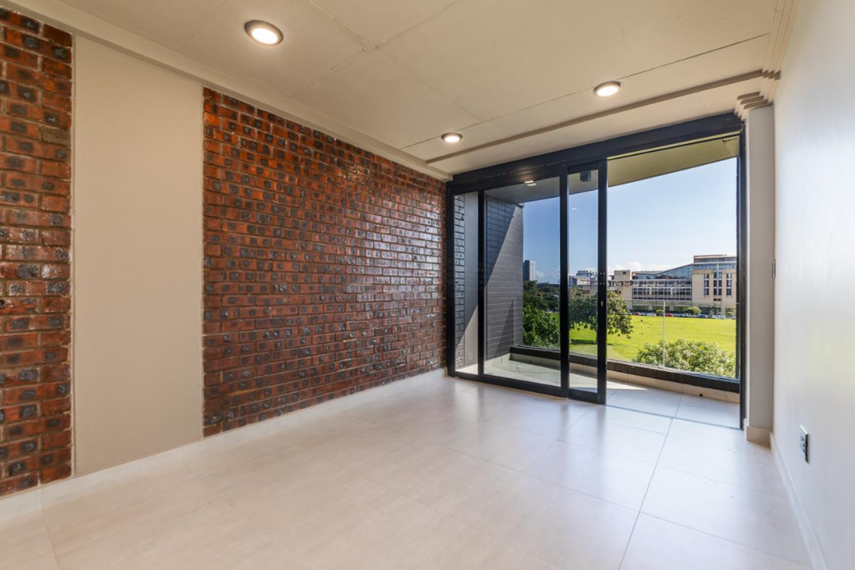 An empty living room featuring a striking exposed brick feature wall, large sliding glass doors, and neutral floor tiles with access to a balcony.