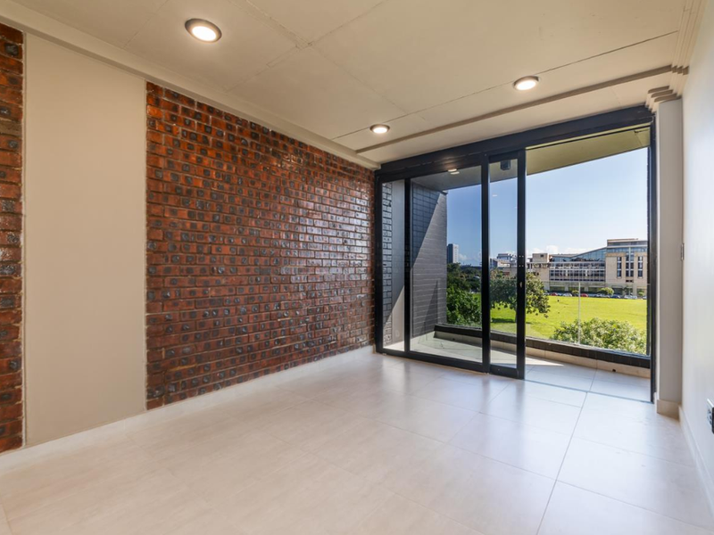 An empty living room featuring a striking exposed brick feature wall, large sliding glass doors, and neutral floor tiles with access to a balcony.