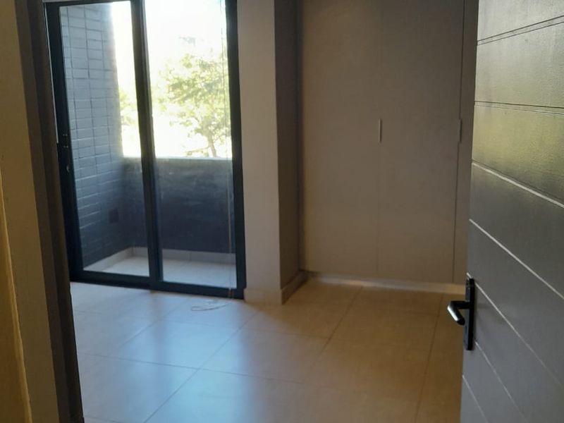 A bright, empty bedroom featuring neutral floor tiles, built-in cupboards, and sliding glass doors leading to a private balcony.