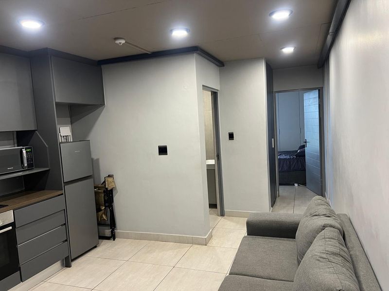 Modern open-plan living area featuring a sleek grey kitchen with built-in cabinetry, tiled flooring, and recessed ceiling lighting, leading to a bedroom and bathroom.