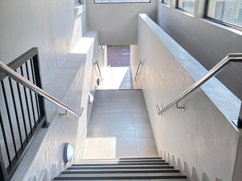 A well-maintained indoor stairwell featuring tiled steps with non-slip strips, stainless steel handrails, and large windows providing natural light.