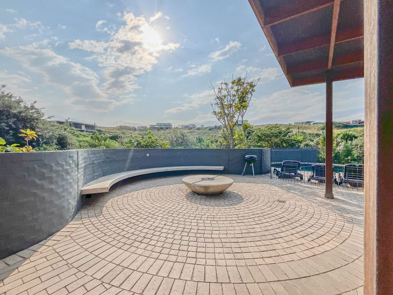 A spacious covered patio featuring a circular paved design, built-in stone bench seating, and a central stone fire pit overlooking the landscape.