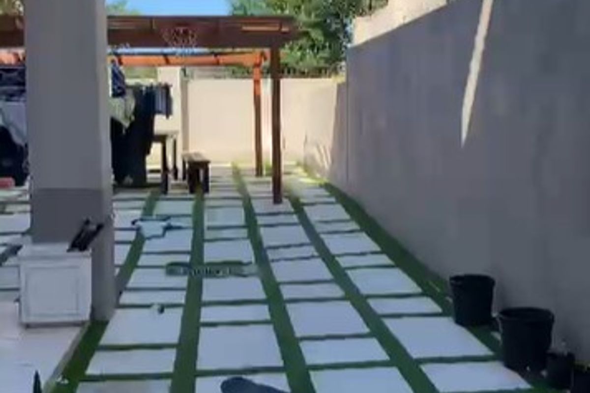 A side courtyard area featuring paved stone tiles with artificial grass infill, a wooden pergola structure, and high perimeter boundary walls.