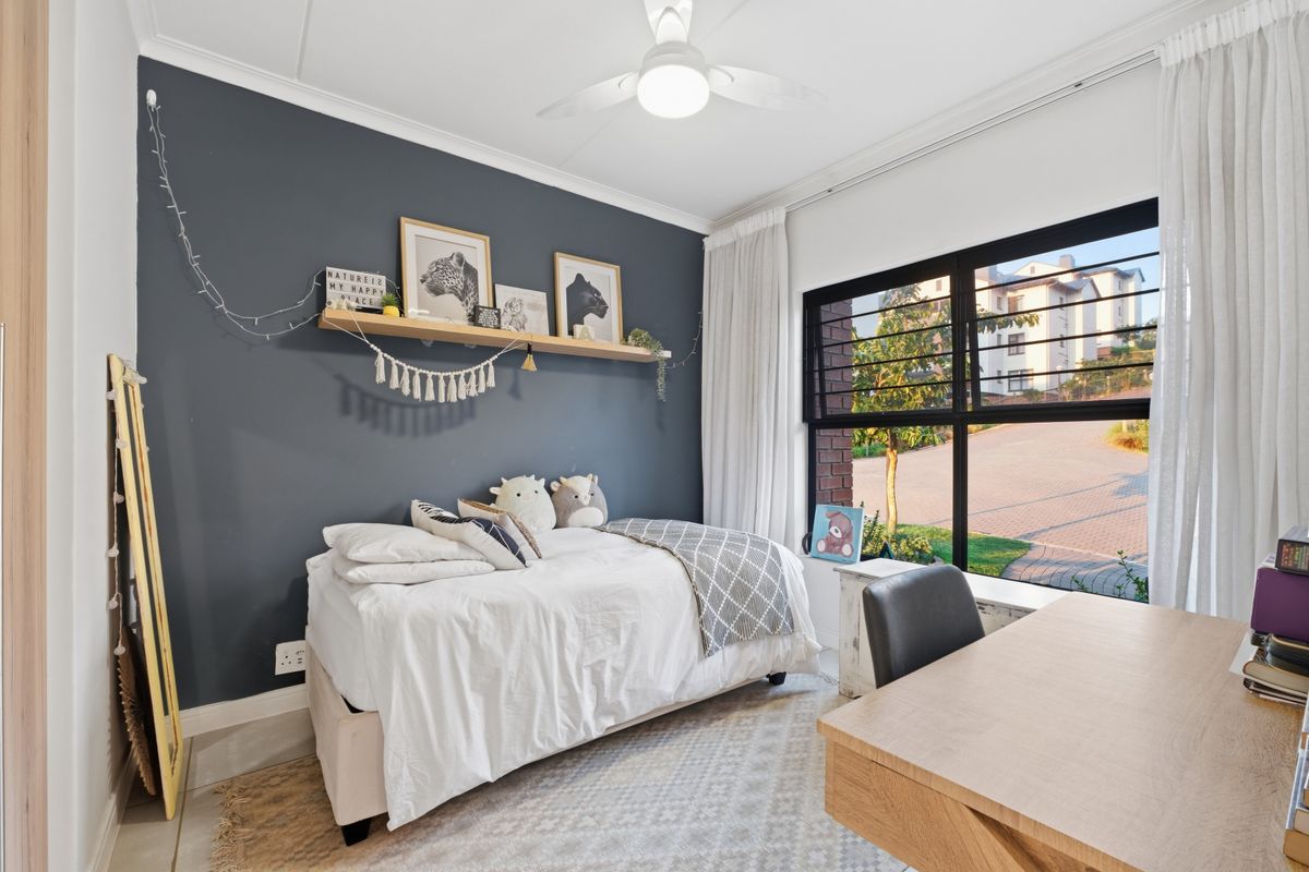 A well-lit bedroom featuring a dark accent wall, a ceiling fan, and large windows with views of the complex driveway.