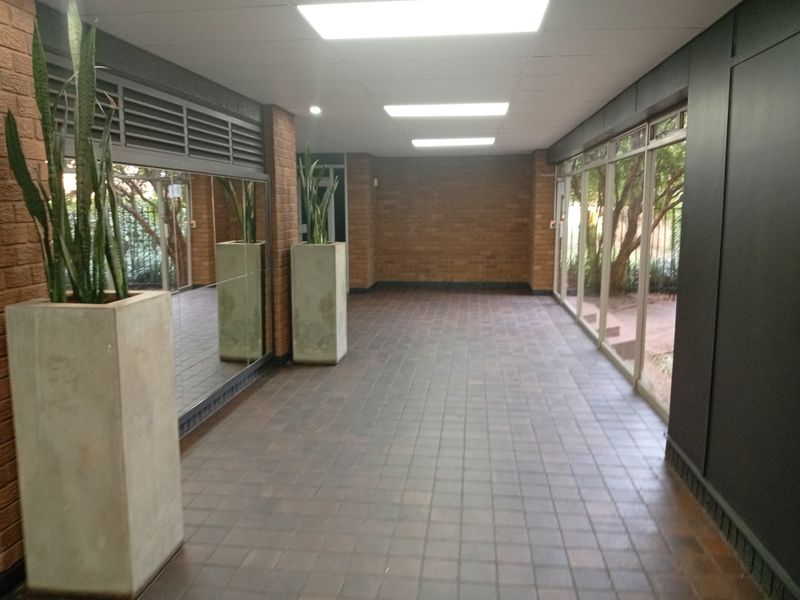 A spacious, well-lit hallway featuring tiled flooring, exposed brick walls, and large windows that provide natural light and views of the surrounding greenery.