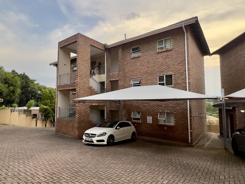 Exterior view of a multi-story face brick apartment building featuring a paved driveway and covered parking bays.