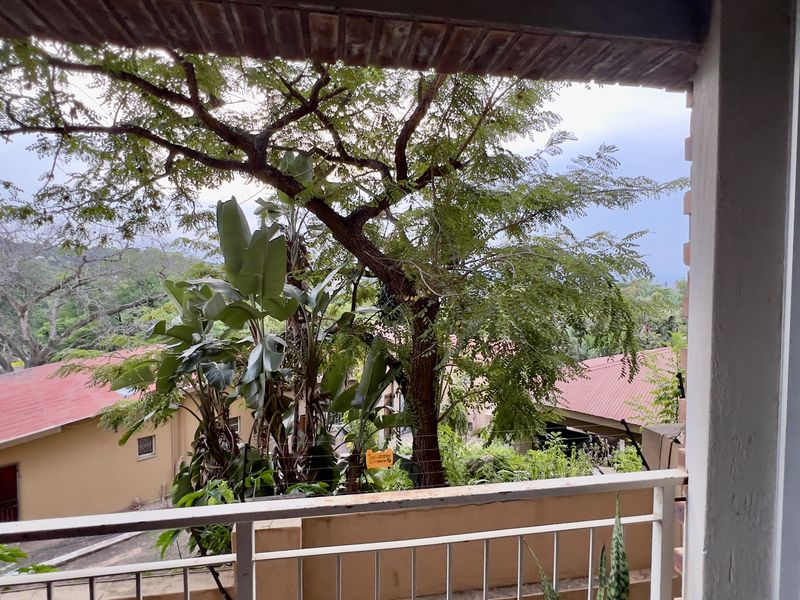 A balcony overlooking a lush garden and surrounding residential properties, featuring a metal balustrade and a concrete ceiling.