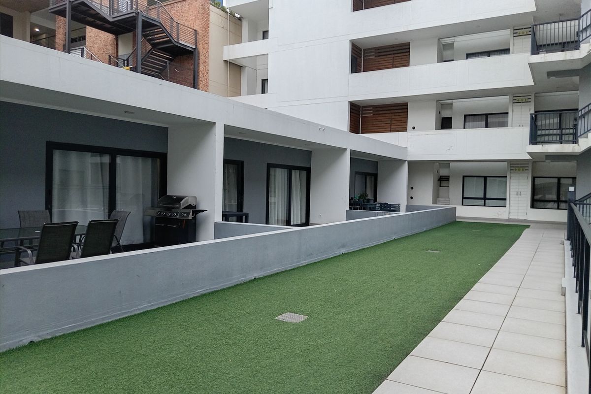 A modern apartment complex featuring a shared outdoor patio area with artificial turf, paved walkways, and covered terrace spaces.