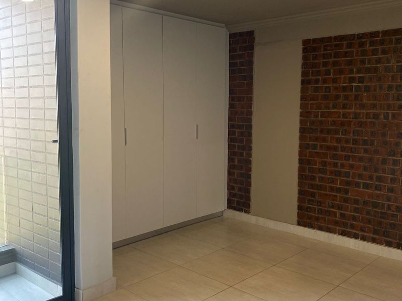 A bright bedroom featuring built-in white cabinetry, exposed brick feature wall, tiled flooring, and sliding glass door access to a balcony.