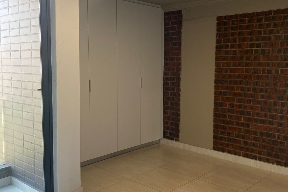 A bright bedroom featuring built-in white cabinetry, exposed brick feature wall, tiled flooring, and sliding glass door access to a balcony.