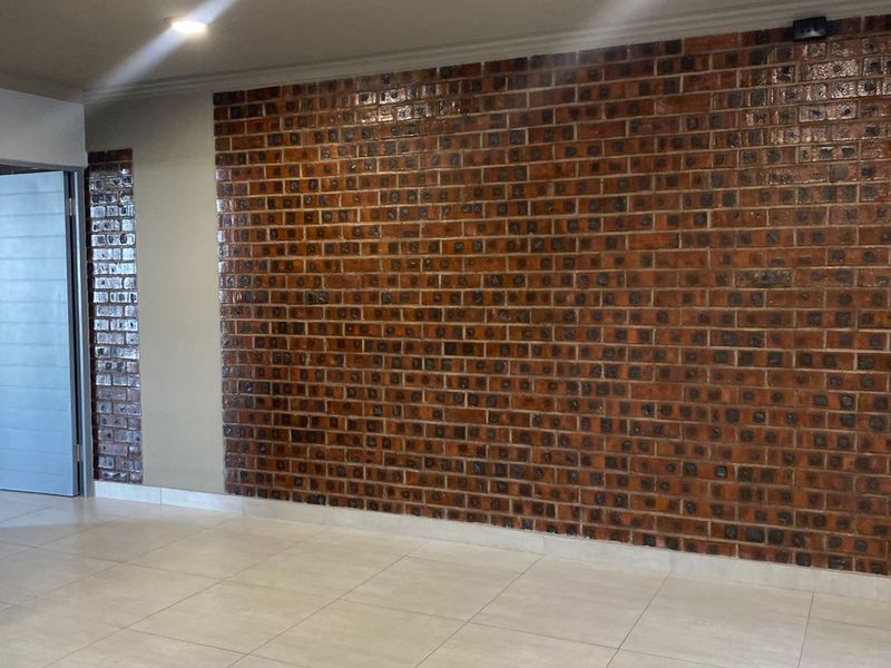 A spacious living area featuring a prominent decorative face-brick feature wall and neutral ceramic floor tiles, illuminated by recessed ceiling downlights.