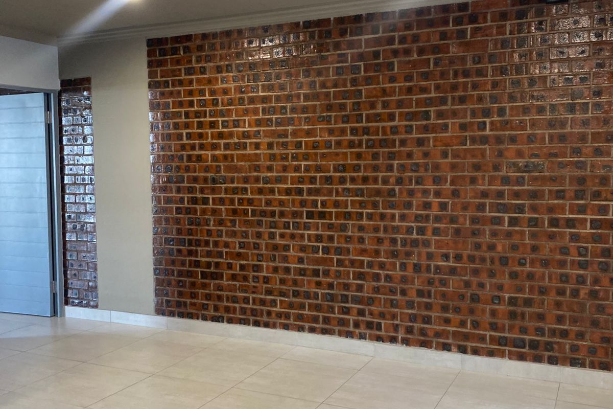 A spacious living area featuring a prominent decorative face-brick feature wall and neutral ceramic floor tiles, illuminated by recessed ceiling downlights.
