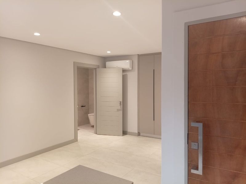 A modern bedroom featuring neutral tiled flooring, built-in cabinetry, recessed ceiling lighting, and an en-suite bathroom with a wall-mounted air conditioning unit.